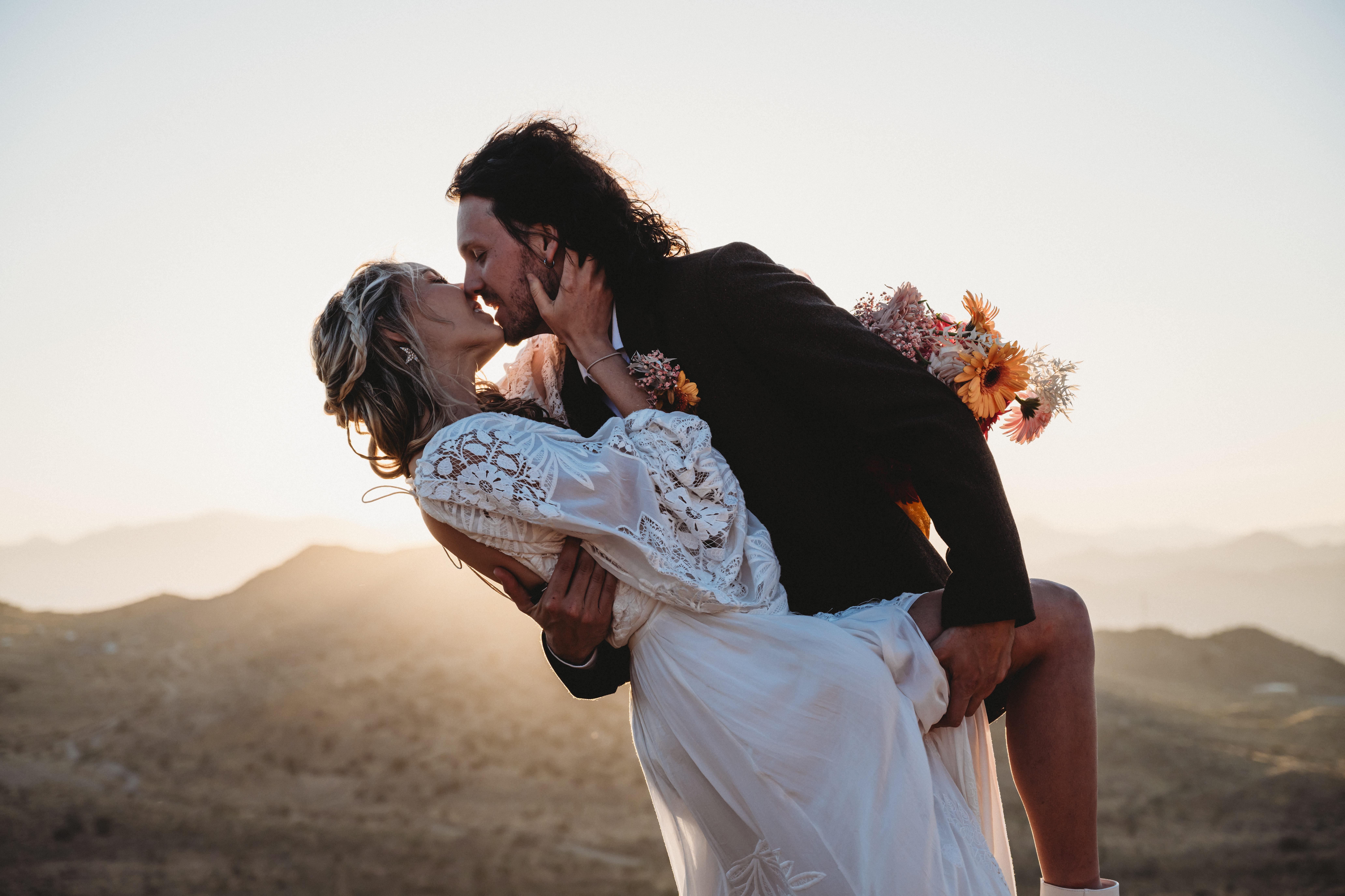 couple kisses at the top of a cliff in Joshua tree national park with the sun setting behind them at their boho elopement